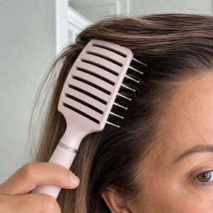 Woman using a pale pink anti-static vent brush at the roots to smooth hair and create lift during styling.