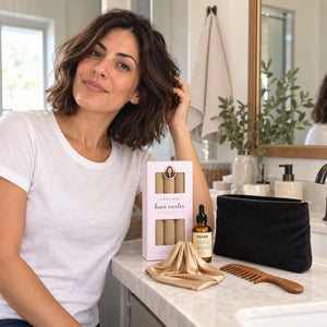 Woman with shoulder-length dark wavy hair showcasing beige heatless curlers, hair serum, black cosmetic pouch, champagne satin bonnet and wooden wide-tooth comb.