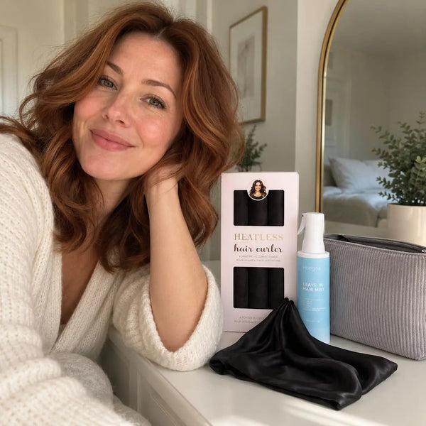 Woman with long auburn wavy hair showcasing black heatless curlers, leave-in hair mist, grey cosmetic pouch and black satin bonnet.
