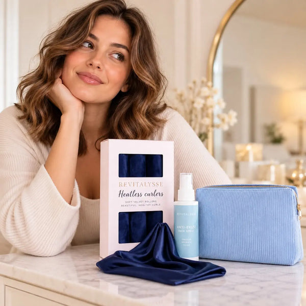 Woman with long chestnut brown wavy hair showcasing Revitalysse navy blue heatless curlers, anti-frizz hair spray, light blue cosmetic pouch and navy satin bonnet.