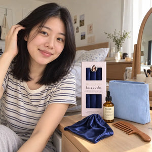 Woman with shoulder-length dark hair showcasing navy blue heatless curlers, hair serum, light blue cosmetic pouch, satin bonnet and wooden wide-tooth comb.