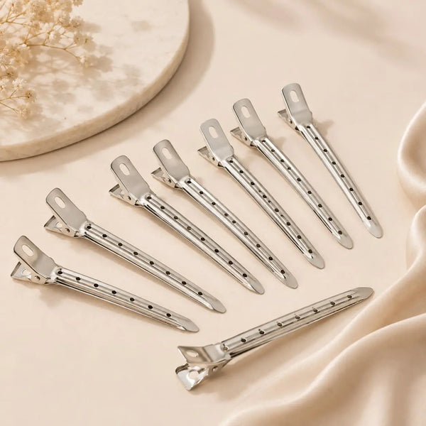 Silver metal hair clips arranged on a neutral satin background for securing volume hair rollers during blowout styling.