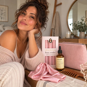 Woman with brunette hair in a messy bun showcasing pink heatless curlers, hair serum, pink cosmetic pouch, pink satin bonnet and wooden wide-tooth comb.