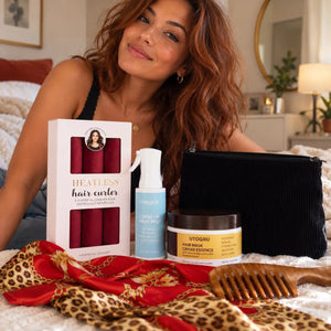 Woman with long auburn wavy hair showcasing red heatless curlers, hair mist, nourishing hair mask, black pouch, silk scarf and wooden comb.
