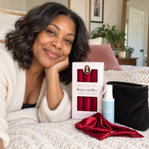 Mature woman with shoulder-length dark wavy hair showcasing red heatless curlers, leave-in hair mist, black cosmetic pouch and red satin bonnet.